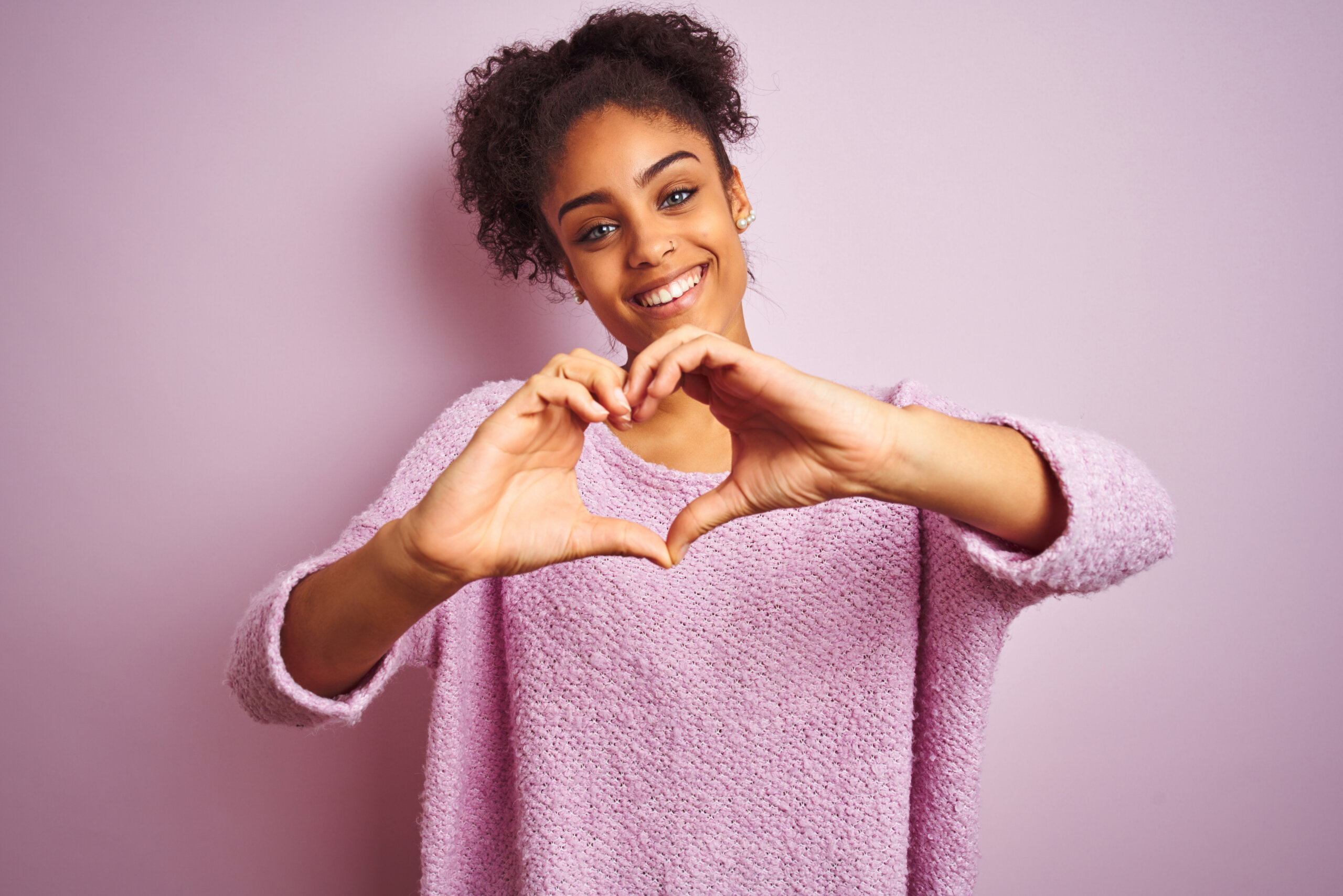 Young Black woman wearing a pink Winter sweater standing in front of a pink background making a heart with her hands
