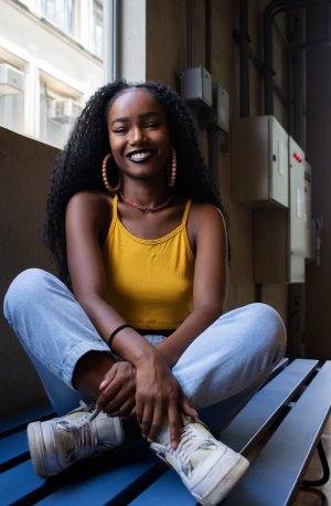 Black Woman wearing a yellow tank top and jeans, smiling with her feet crossed
